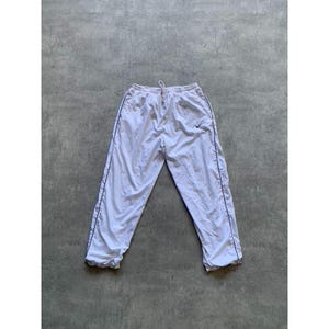 May include: White Nike track pants with a drawstring waist and black trim along the sides. The Nike swoosh logo is visible on the left leg. The pants are made of a lightweight material.