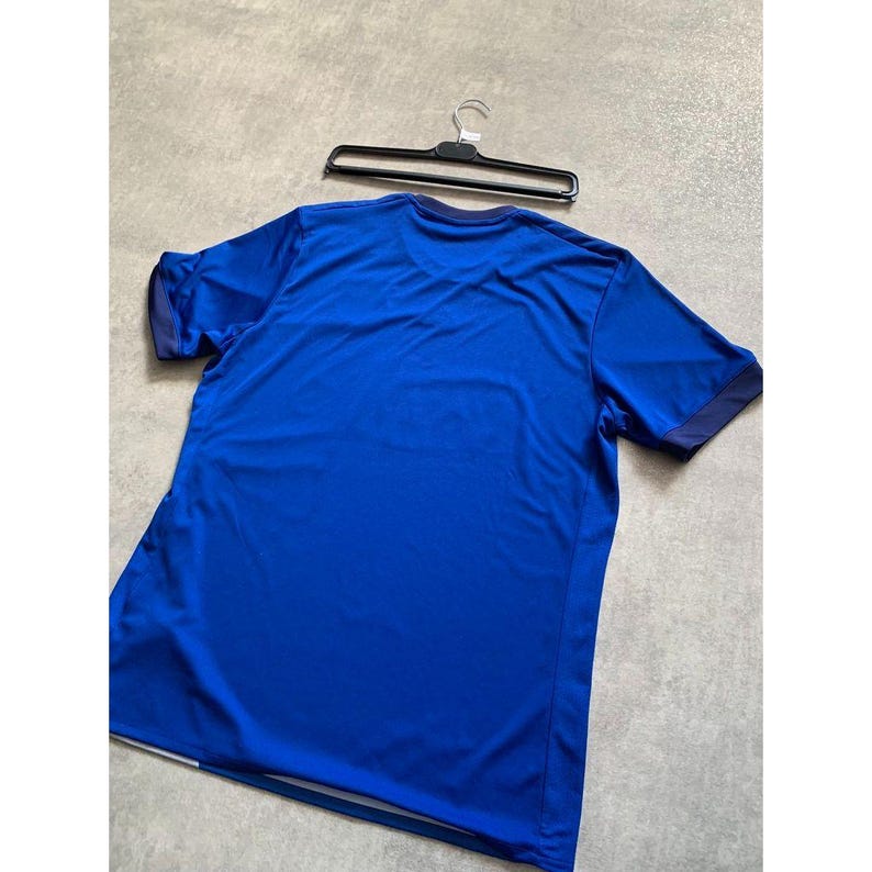 Pu&ograve; includere: Una maglietta sportiva blu reale a maniche corte con finiture blu navy sulle maniche e sul colletto. La maglietta &egrave; appesa a una gruccia nera. Realizzata in materiale leggero e traspirante.