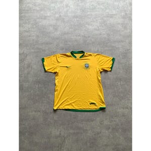 May include: A yellow soccer jersey with green trim on the collar and sleeves. The jersey features a small black Nike swoosh and a crest with a blue and green design. The jersey is laid flat on a gray surface.