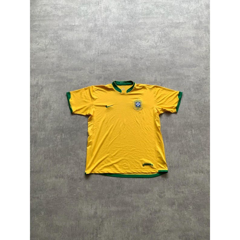 May include: A yellow soccer jersey with green trim on the collar and sleeves. The jersey features a small black Nike swoosh and a crest with a blue and green design. The jersey is laid flat on a gray surface.