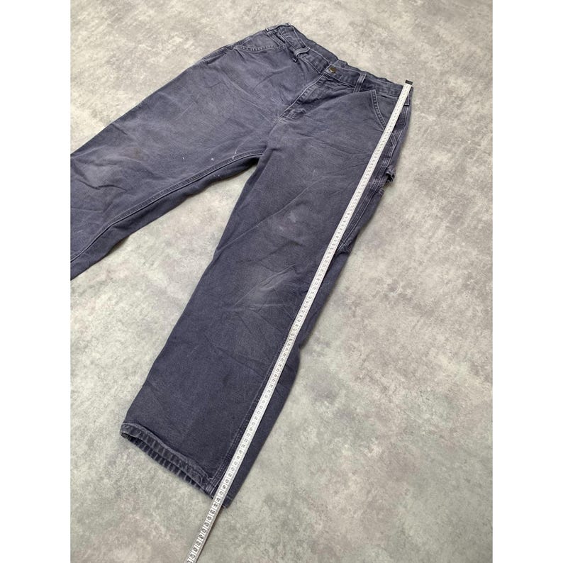 May include: Faded navy blue work pants with a straight leg cut. A white measuring tape is laid along the length of the pants. The pants have multiple pockets and a button and zipper closure. The fabric shows signs of wear.