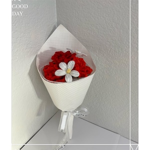 May include: A bouquet of red flowers with a white flower accent, wrapped in white paper and tied with a ribbon. The bouquet is presented in a corner, with the text "HAVE A GOOD DAY" visible in the upper left corner.