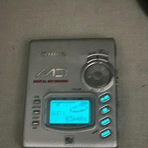 Könnte beinhalten: Silbernes MD-Digitalaufnahmegerät mit blau beleuchtetem Display. Das Gerät hat einen Lautstärkeregler, mehrere Tasten und einen kleinen Bildschirm mit Informationen. Der Text "MD DIGITAL RECORDING" ist sichtbar.