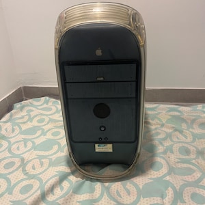 May include: A vintage Apple iMac G3 computer in a translucent blue case. The computer has a dark grey front panel with a built-in CD-ROM drive and speaker. The Apple logo is visible on the front. The computer is sitting on a patterned bedspread.