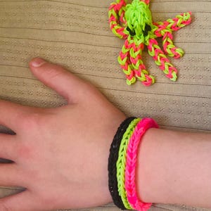 May include: A neon green and pink rubber band octopus figure and three rubber band bracelets. The bracelets are black, neon green, and pink. The octopus has black eyes and is on a tan surface.