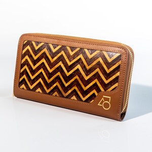 Può includere: Un portafoglio in pelle marrone con un motivo a zig-zag in legno chiaro e scuro. Il portafoglio ha una cerniera dorata e un logo dorato sul davanti. Il portafoglio è rettangolare e sembra nuovo.