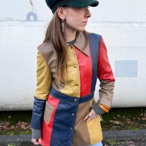 May include: A patchwork leather jacket in brown, yellow, red, and blue. The jacket has a collared neckline and button closures. A dark green corduroy cap is also visible. The jacket's design is reminiscent of the 1970s.