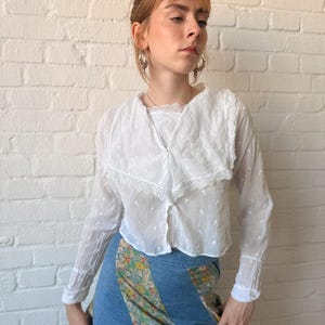 May include: A white long-sleeved blouse featuring a large collar with lace trim and a subtle swiss dot texture, paired with a blue denim skirt that has diagonal panels of floral fabric in shades of yellow, green, and white.