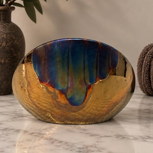 May include: A semi-circular decorative bowl featuring a hammered gold metallic finish on the lower portion and a smooth, iridescent blue glaze dripping down from the top edge, creating a distinct contrast between the two textures and colors.