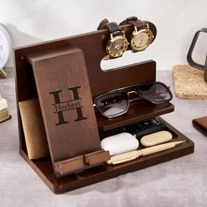 Könnte beinhalten: Ein dunkelbrauner Holz-Organizer mit Fächern für Uhren, Sonnenbrillen, ein Telefon und anderes Zubehör. Der Organizer trägt den Namen "Herbert" auf der Vorderseite eingraviert. Zwei Uhren und eine Sonnenbrille sind ausgestellt.