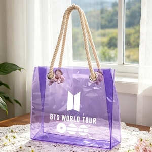 Puede incluir: Un bolso tote transparente de color púrpura con asas de cuerda. La bolsa presenta el texto "BTS WORLD TOUR" en blanco, junto con un logotipo. La bolsa está sobre una mesa de madera con un tapete de encaje blanco.
