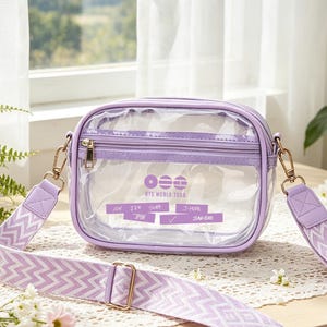 以下が含まれることがあります： ラベンダー色のトリムとストラップが付いた、透明な長方形のクロスボディバッグ。バッグはジッパー開閉式で、ゴールドカラーの金具が付いています。ストラップは、白いシェブロン柄のラベンダー色です。バッグには「BTS WORLD TOUR」と書かれています。