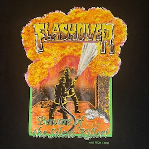 Può includere: T-shirt nera con una grafica raffigurante due vigili del fuoco che combattono un incendio. Il design include il testo "FLASHOVER" in arancione e giallo, e la frase "Beware of the Silent Killer!" in verde. La grafica è del 1992.