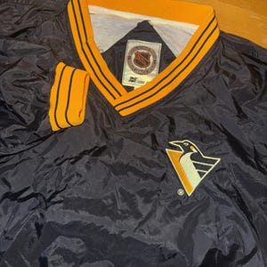 May include: Black and gold NHL Pittsburgh Penguins pullover jacket. The jacket features a V-neck collar and sleeve cuffs with gold and black stripes. The Penguins logo is on the lower right. The NHL logo is on the tag.