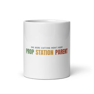 Può includere: Una tazza in ceramica bianca con il testo "ONE MORE CUTTING WON'T HURT" in piccole lettere nere sopra il testo più grande e multicolore "PROP STATION PARENT" in verde, giallo e rosso, centrato sulla parte anteriore del contenitore cilindrico.