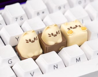 Könnte beinhalten: Eine weiße mechanische Tastatur mit individuellen Katzen-Tastenkappen. Die Tastenkappen sind hellgelb mit braunen und orangefarbenen Details und haben die Form von Cartoon-Katzen. Die Tastatur hat weiße Tasten und einen schwarzen Hintergrund.