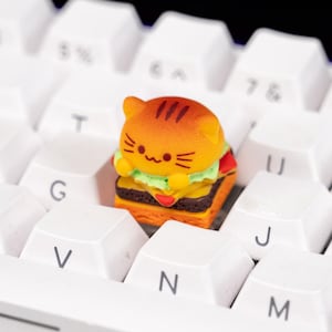 May include: A novelty keycap featuring a cartoon cat design in the shape of a hamburger. The keycap is orange, yellow, and brown, and sits atop a white keyboard. The keyboard has white keys with black lettering.