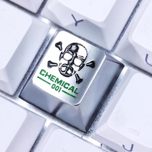 Op de afbeelding: Close-up van een toetsenbord met witte toetsen. Eén toets is een aangepast ontwerp met een schedel- en kruisbogensymbool en het woord "CHEMICAL" in het groen. De toets is zwart met een zilveren bovenkant.