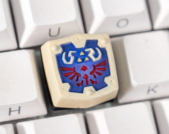 May include: A custom keycap featuring a shield design with blue, red, and white accents, set against a white keyboard. The keycap is a unique replacement for a standard keyboard key, adding a personalized touch to the setup.