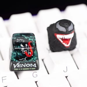 May include: Two custom keycaps featuring the Venom character from Marvel Comics. One keycap has a black and green design with the word "Venom". The other keycap is black with a white and red Venom face.