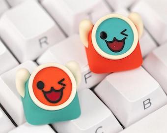 May include: Two colorful custom keycaps with a cartoon drum design, featuring a winking face and a playful expression. The keycaps are red, teal, and orange, with white accents, resting on a white keyboard.