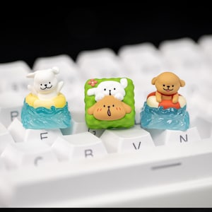 May include: Three custom keycaps featuring cartoon dog designs. One dog is in a yellow and white inner tube, another is on a green square, and the third is in an orange life preserver. The keycaps are on a white keyboard.