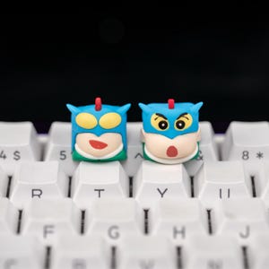 May include: Two novelty keycaps with cartoon character designs. The keycaps are blue with yellow, red, and white accents, and are placed on a white keyboard. The keycaps have a superhero theme.