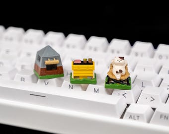 May include: Miniature keyboard keycaps featuring a camping theme. Includes a tent, a yellow barbecue grill, and a cat relaxing in a chair. The keycaps are in various colors and sit on a white keyboard.