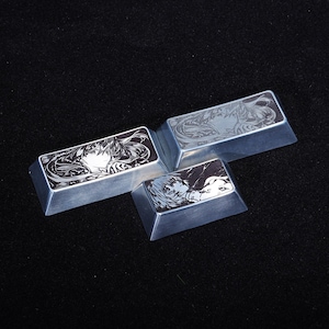 May include: Three silver-colored rectangular keycaps with anime-style character designs. Each keycap features a different black and white illustration. The keycaps are arranged on a dark surface.