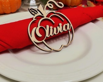 Puede incluir: Un tarjetero de mesa de madera con forma de manzana con el nombre "Olivia" grabado, colocado sobre una servilleta roja y un plato blanco. El entorno incluye calabazas y follaje otoñal, con un tema de otoño.