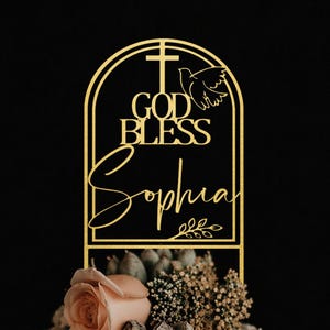 May include: A two-tiered white cake decorated with flowers and greenery, topped with a gold cake topper. The topper features a cross, dove, and the words "GOD BLESS Sophia" in elegant script, perfect for a baptism or christening.