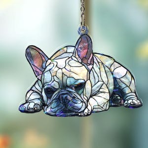 May include: A decorative hanging ornament featuring a French Bulldog in a stained-glass style. The dog is depicted lying down, with a mosaic pattern in shades of white, blue, and purple. The ornament hangs from a silver chain.