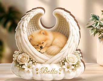 May include: A wooden angel wings decoration with a sleeping Pomeranian dog inside. The wings are white with brown edges, and the dog is light brown. The base features flowers and the name "Bella" in gold script.