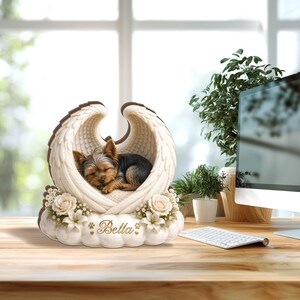 May include: A memorial figurine featuring a sleeping Yorkshire Terrier nestled within angel wings. The wings are cream-colored with brown trim, resting on a cloud-shaped base adorned with flowers. The name "Bella" is inscribed on the base.