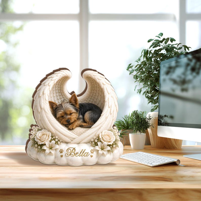 May include: A memorial figurine featuring a sleeping Yorkshire Terrier nestled within angel wings. The wings are cream-colored with brown trim, resting on a cloud-shaped base adorned with flowers. The name "Bella" is inscribed on the base.