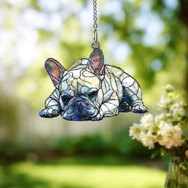 May include: A decorative hanging ornament featuring a French Bulldog in a stained glass style. The dog is lying down, with a detailed pattern of white, blue, and purple hues. The ornament hangs from a silver chain, perfect for a pet lover.