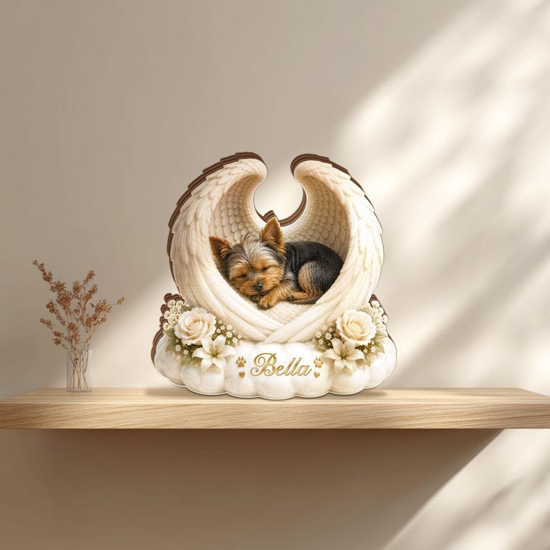 May include: A decorative memorial featuring a sleeping Yorkshire Terrier inside angel wings. The wings are white with brown trim, resting on a cloud-shaped base with floral accents. The name "Bella" is inscribed in gold.