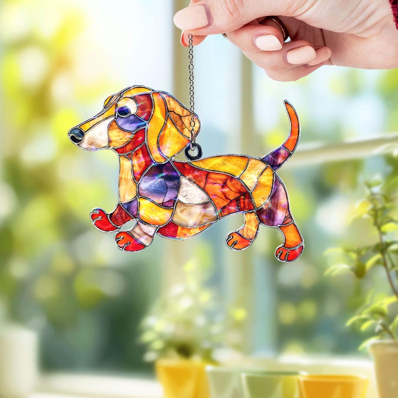 May include: Stained glass dachshund dog suncatcher with a chain for hanging. The dog features a colorful mosaic design with shades of orange, yellow, red, and purple. The dog's paws and tail are red.