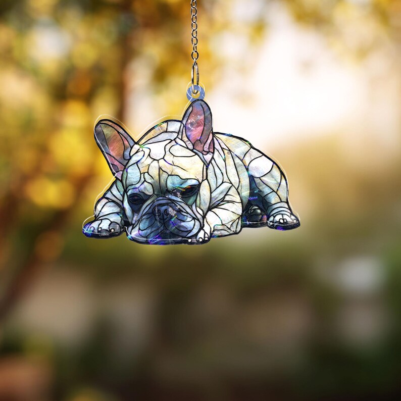 May include: A decorative hanging ornament featuring a French Bulldog in a stained-glass style. The dog is depicted lying down, with a detailed pattern of white, blue, and purple hues. The ornament hangs from a silver chain, perfect for pet lovers.