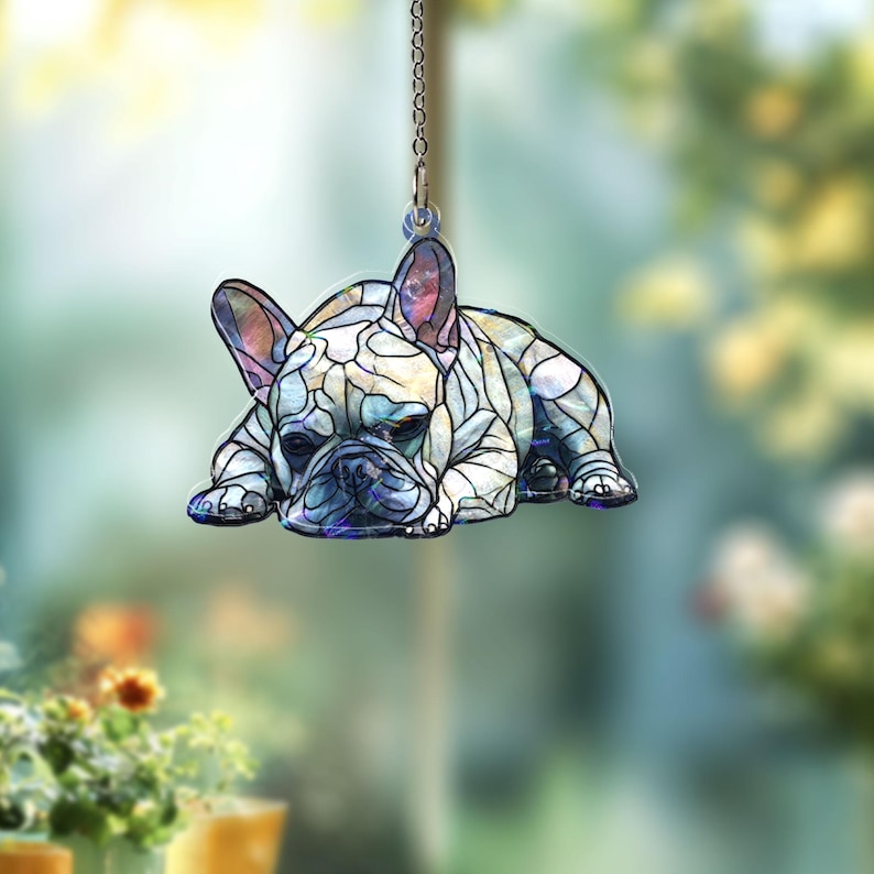 May include: A decorative hanging ornament featuring a French Bulldog in a stained-glass style. The dog is depicted lying down, with a mosaic pattern in shades of white, blue, and purple. The ornament hangs from a silver chain.