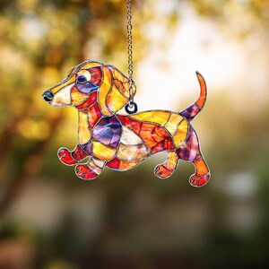 May include: A stained glass dachshund dog ornament. The dog is made of colorful glass pieces in shades of orange, yellow, red, and purple. It is hanging from a silver chain. The dog is in a running pose, with its tail curved upwards.