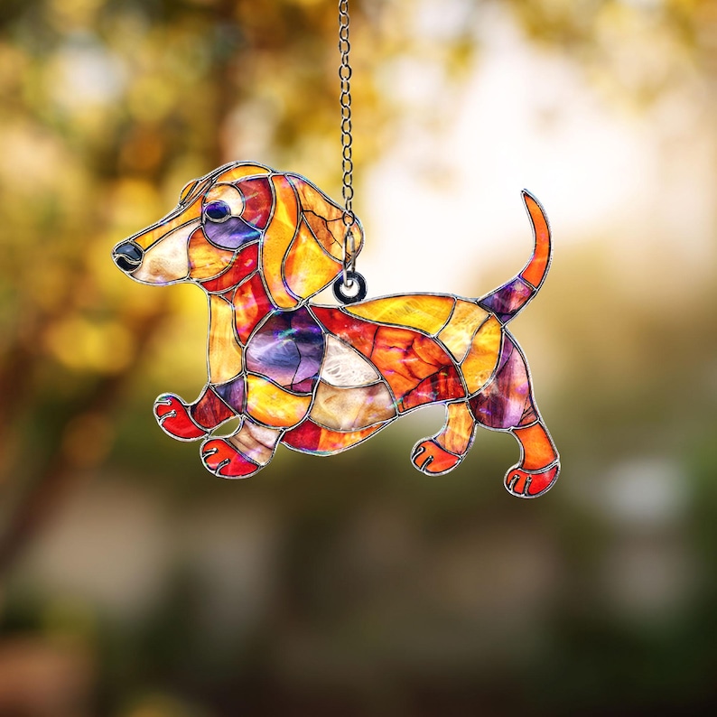 May include: A stained glass dachshund dog ornament. The dog is made of colorful glass pieces in shades of orange, yellow, red, and purple. It is hanging from a silver chain. The dog is in a running pose, with its tail curved upwards.