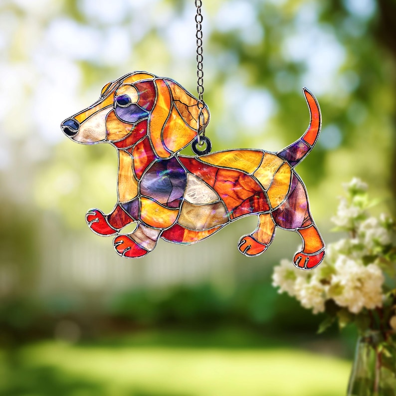 May include: Stained glass dachshund dog suncatcher with a chain for hanging. The dog features a patchwork design in shades of orange, yellow, red, and purple. The dog is in a running pose, with its tail raised.
