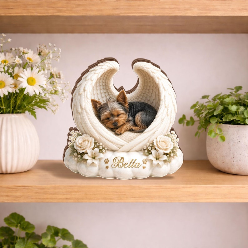 May include: A decorative memorial featuring a sleeping Yorkshire Terrier nestled within white angel wings. The base is adorned with flowers and the name "Bella" in gold script. The wings have a brown outline.