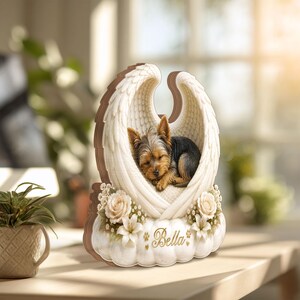 May include: A wooden memorial featuring a sleeping Yorkshire Terrier nestled in angel wings. The wings are white with a brown border, and the base is adorned with flowers and the name "Bella".
