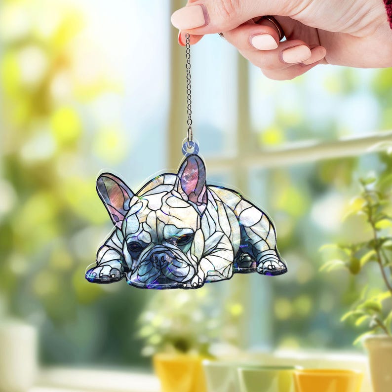 May include: A decorative ornament featuring a French Bulldog in a stained-glass style. The dog is lying down, with a detailed pattern of white, blue, and purple hues. The ornament is suspended by a silver chain, perfect for hanging.