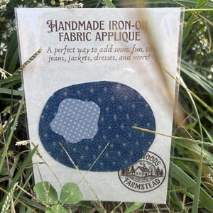 May include: An oval-shaped dark blue fabric applique with a small floral pattern and a light blue scalloped patch overlay. The packaging features the text HANDMADE IRON-ON FABRIC APPLIQUE and a circular logo with a house and the text FARMSTEAD.