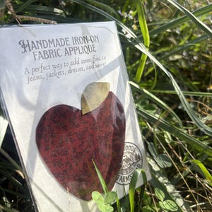 May include: A handmade iron-on fabric applique in the shape of an apple with a dark red floral pattern and a gold leaf. The packaging features the text HANDMADE IRON-ON FABRIC APPLIQUE and a circular logo with the words WOODS FARMS.