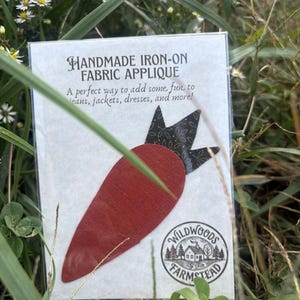 May include: A carrot-shaped iron-on fabric applique featuring a solid rust-colored body and a black top with a white dotted pattern. The applique is packaged on a white card labeled HANDMADE IRON-ON FABRIC APPLIQUE with a Wildwoods Farmstead logo.