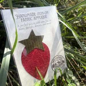May include: A handmade iron-on fabric applique shaped like a strawberry with a red textured body and a dark green star-shaped top. The packaging features the text HANDMADE IRON-ON FABRIC APPLIQUE and a circular logo for WILDWOODS FARMSTEAD.